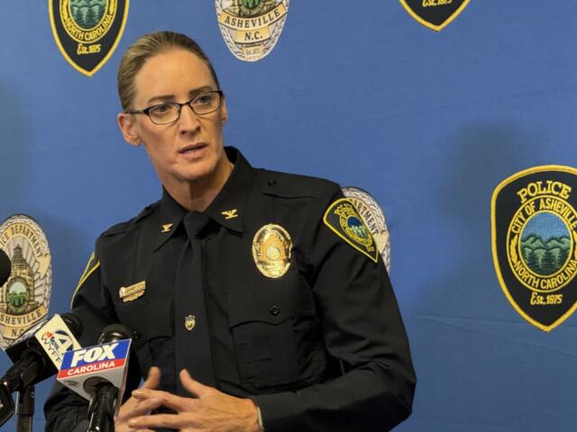 Female police officer in uniform speaks at a press conference, with microphones in front and a blue backdrop dotted with Asheville Police Department patches.
