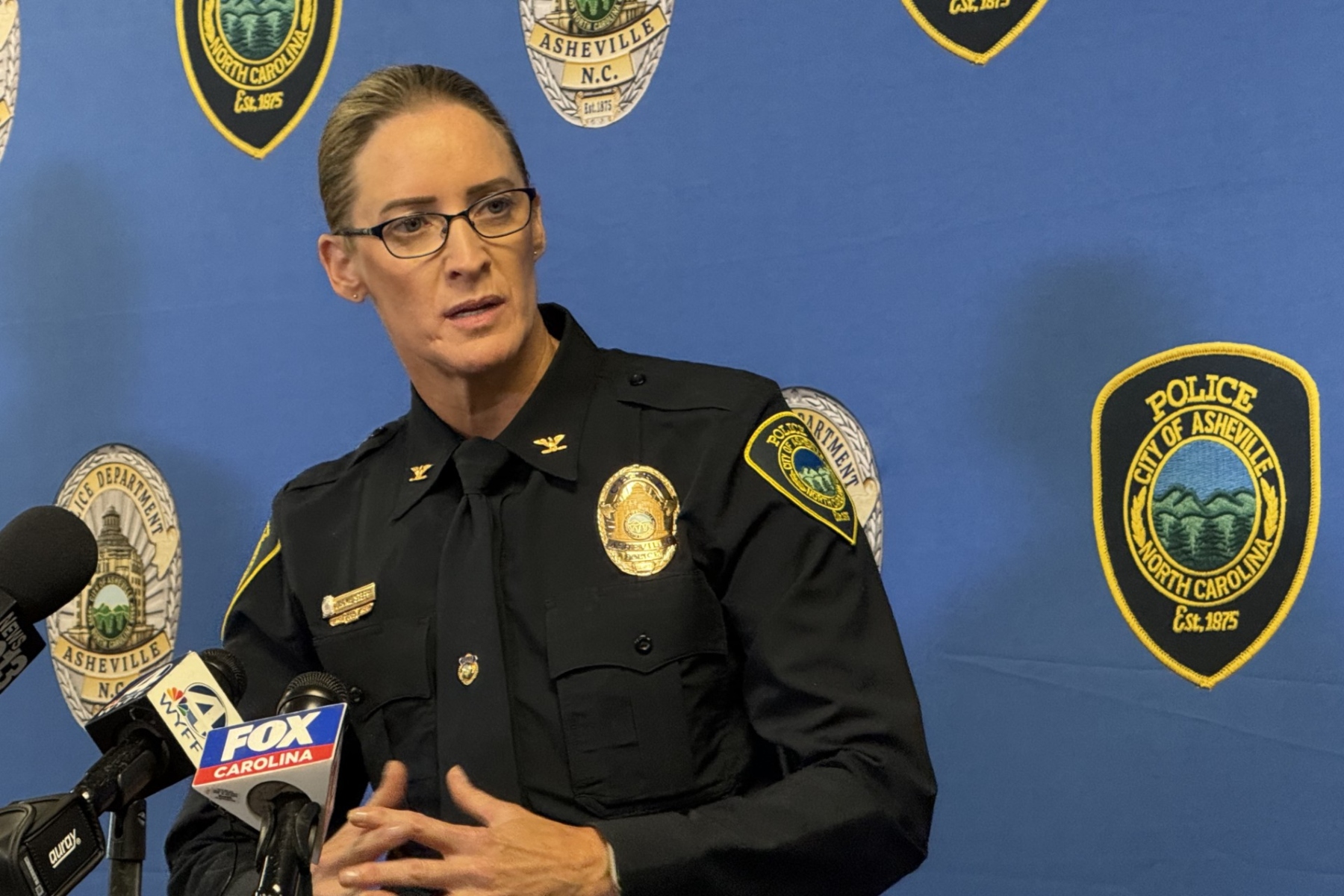 Female police officer in uniform speaks at a press conference, with microphones in front and a blue backdrop dotted with Asheville Police Department patches.