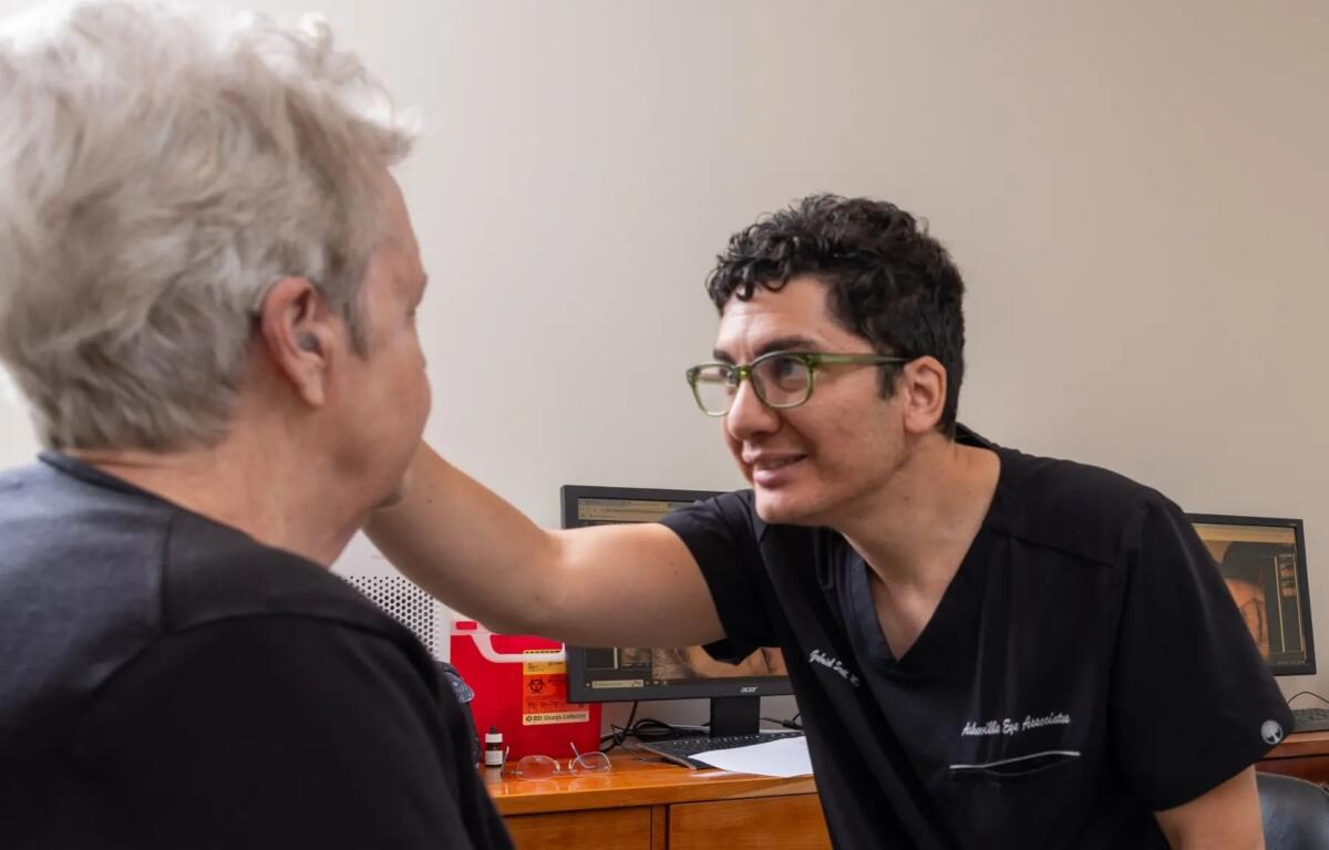 Clinician with green glasses leans toward a patient, smiling during a medical consultation in a clinic office on a wooden desk with monitors in the background.