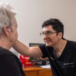 Clinician with green glasses leans toward a patient, smiling during a medical consultation in a clinic office on a wooden desk with monitors in the background.