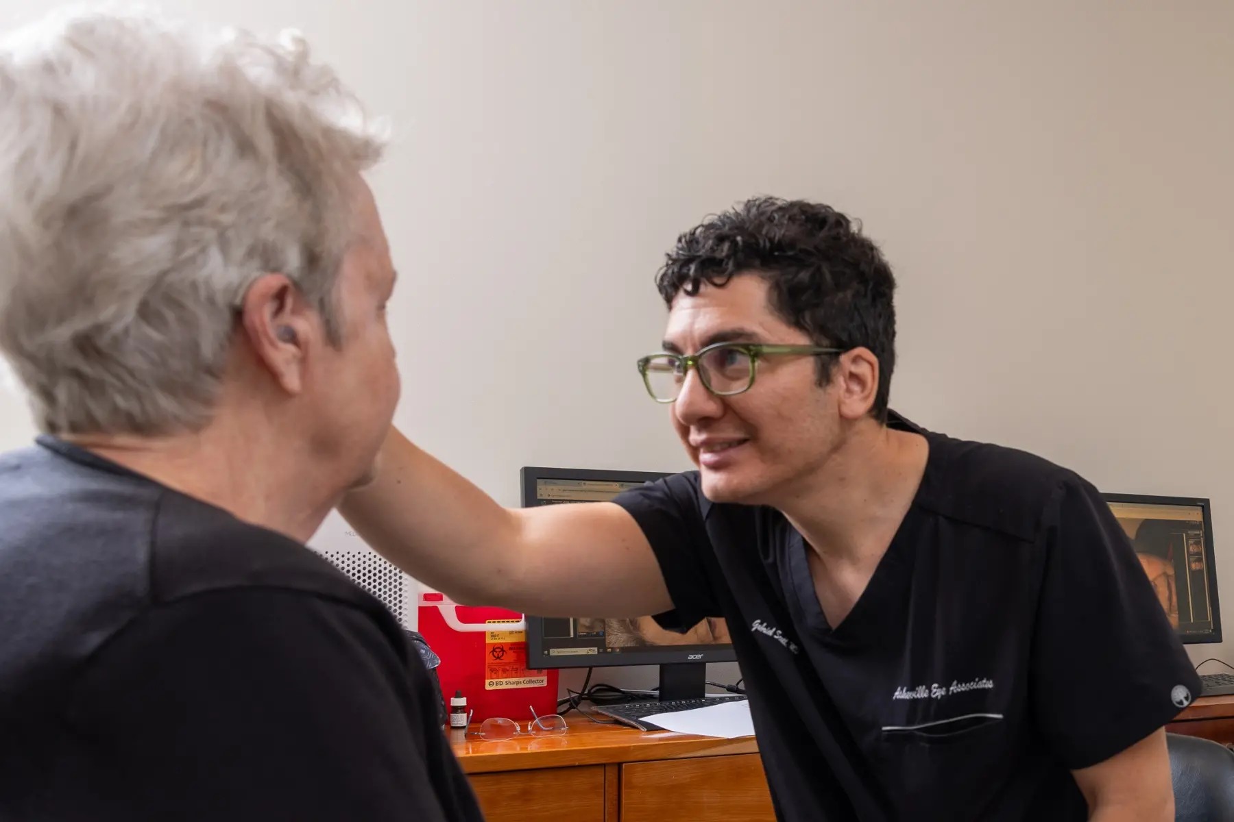 Clinician with green glasses leans toward a patient, smiling during a medical consultation in a clinic office on a wooden desk with monitors in the background.