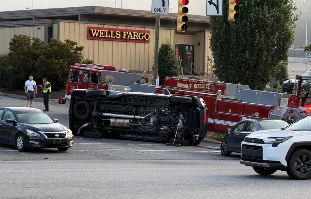 Overturned fire-truck on its side in a parking lot near a Wells Fargo, with firefighters and a police officer nearby.