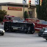 Overturned fire-truck on its side in a parking lot near a Wells Fargo, with firefighters and a police officer nearby.