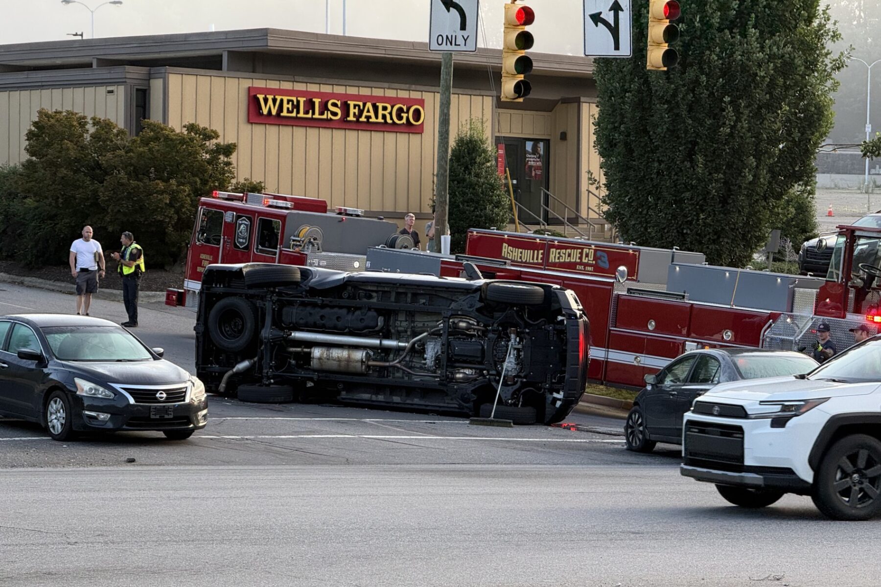 Overturned fire-truck on its side in a parking lot near a Wells Fargo, with firefighters and a police officer nearby.
