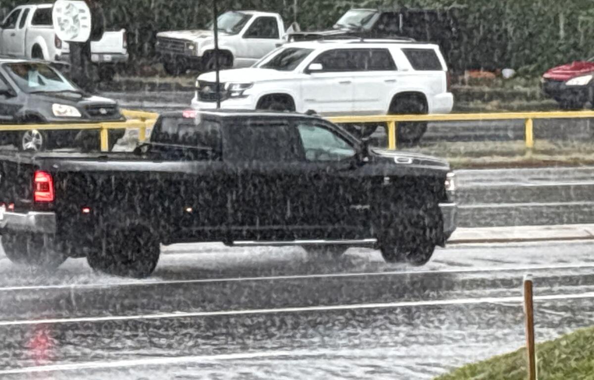 Black pickup truck drives on a wet highway in heavy rain, with yellow guardrails in the background and other vehicles nearby