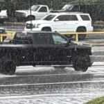 Black pickup truck drives on a wet highway in heavy rain, with yellow guardrails in the background and other vehicles nearby
