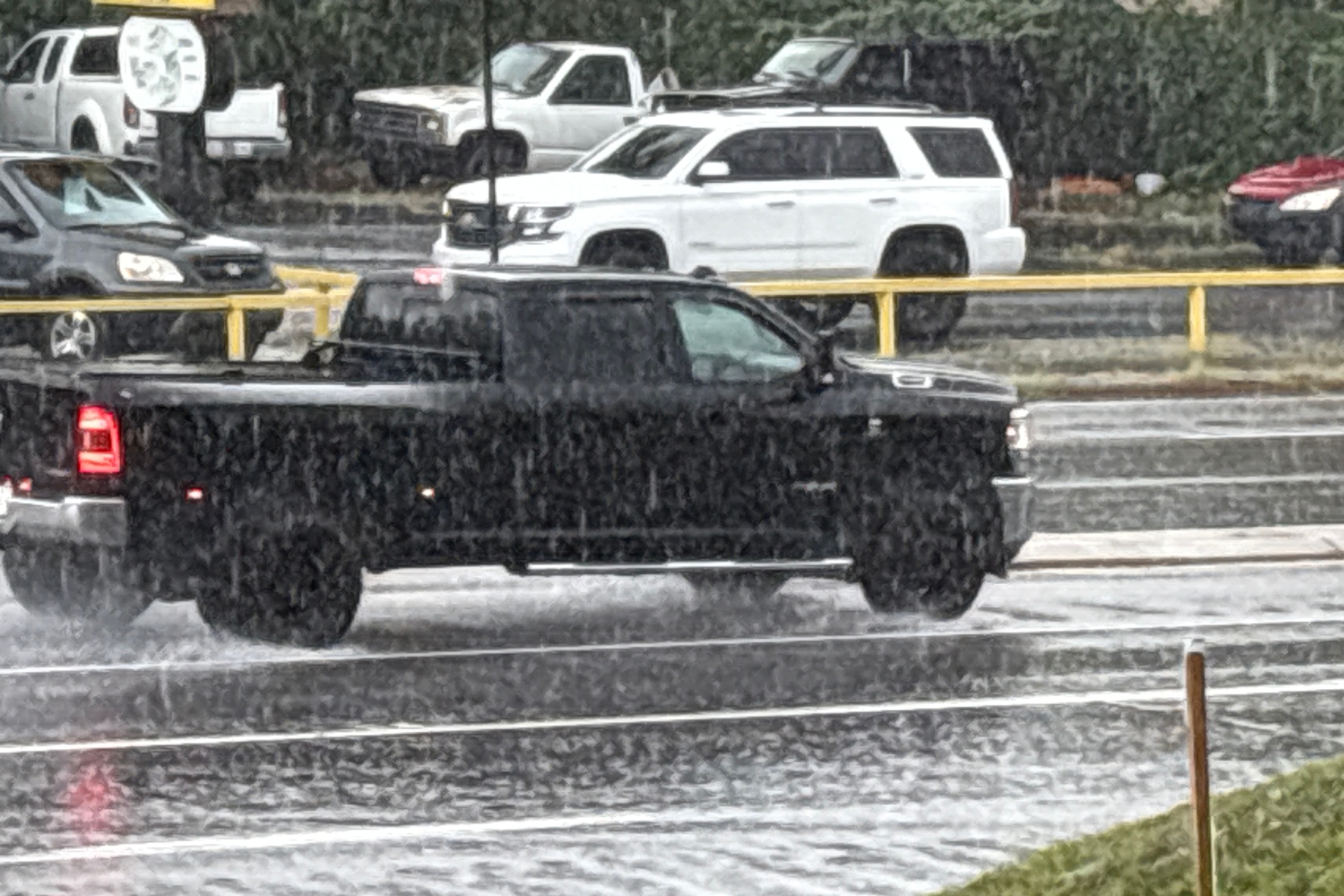 Black pickup truck drives on a wet highway in heavy rain, with yellow guardrails in the background and other vehicles nearby