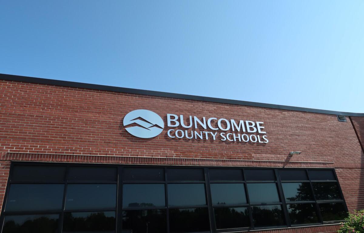 Brick school building with Buncombe County Schools sign and circular logo against a clear blue sky