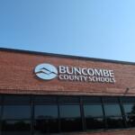 Brick school building with Buncombe County Schools sign and circular logo against a clear blue sky