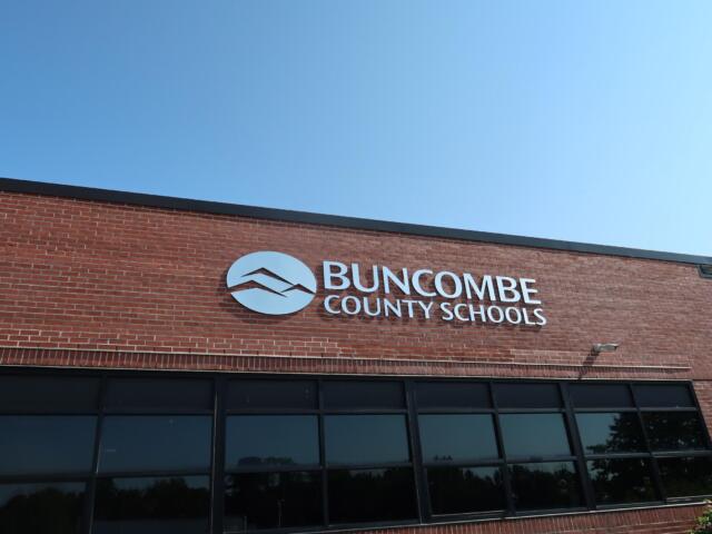 Brick school building with Buncombe County Schools sign and circular logo against a clear blue sky