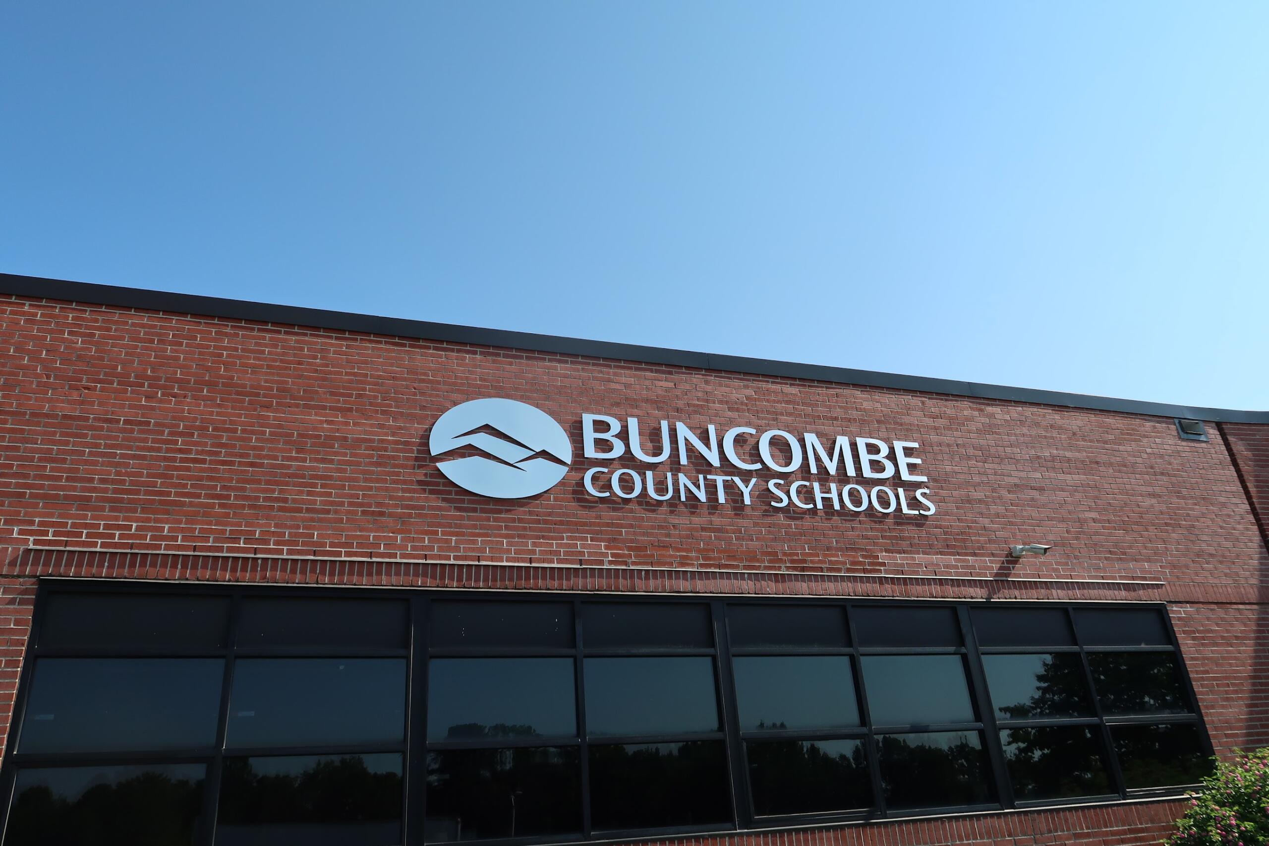 Brick school building with Buncombe County Schools sign and circular logo against a clear blue sky