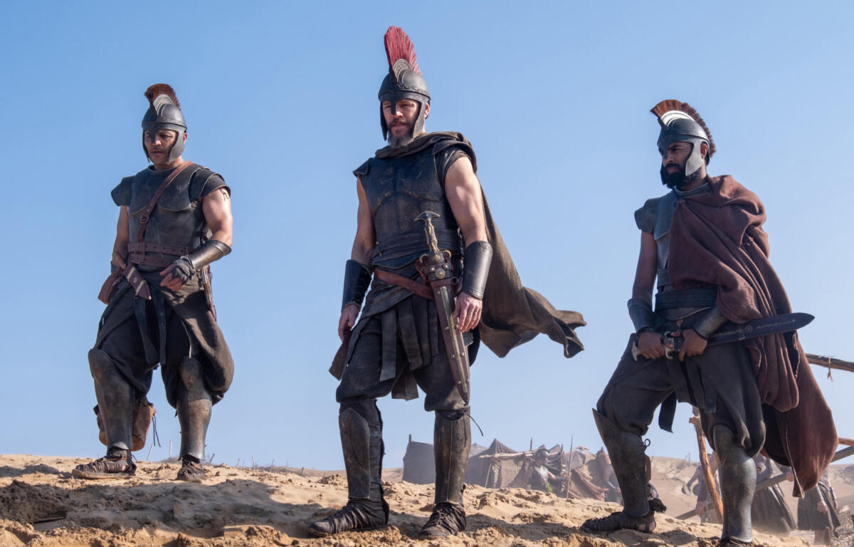 Three ancient warriors in leather and metal armor with plumed helmets stand on a sandy hill against a clear blue sky, with the central figure wearing a red crest.