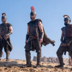Three ancient warriors in leather and metal armor with plumed helmets stand on a sandy hill against a clear blue sky, with the central figure wearing a red crest.