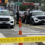 Police cars with flashing blue lights at a taped-off intersection, officers talking near the curb under a 'Short Coxe Ave' sign.