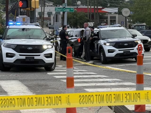 Police cars with flashing blue lights at a taped-off intersection, officers talking near the curb under a 'Short Coxe Ave' sign.