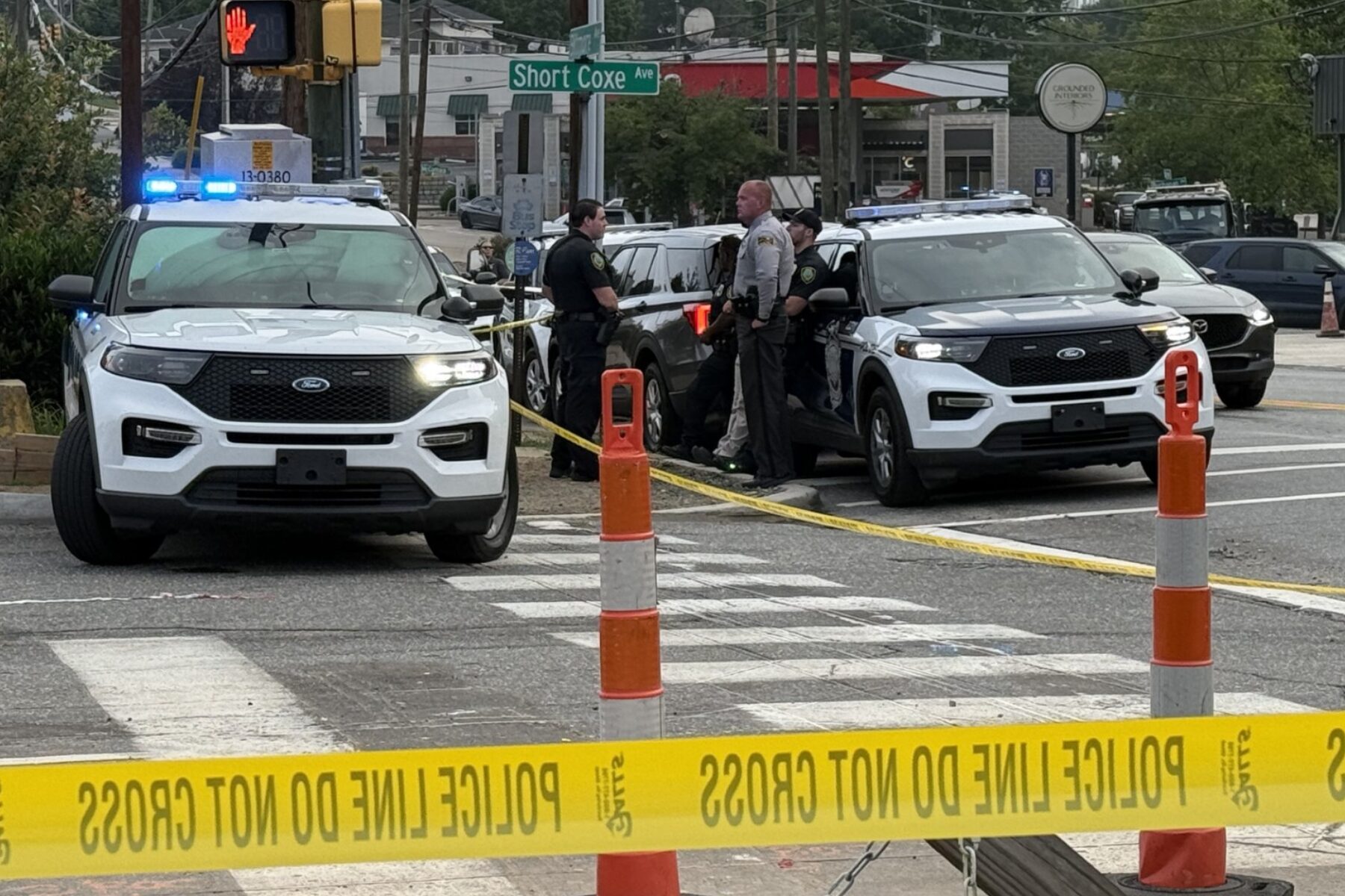 Police cars with flashing blue lights at a taped-off intersection, officers talking near the curb under a 'Short Coxe Ave' sign.