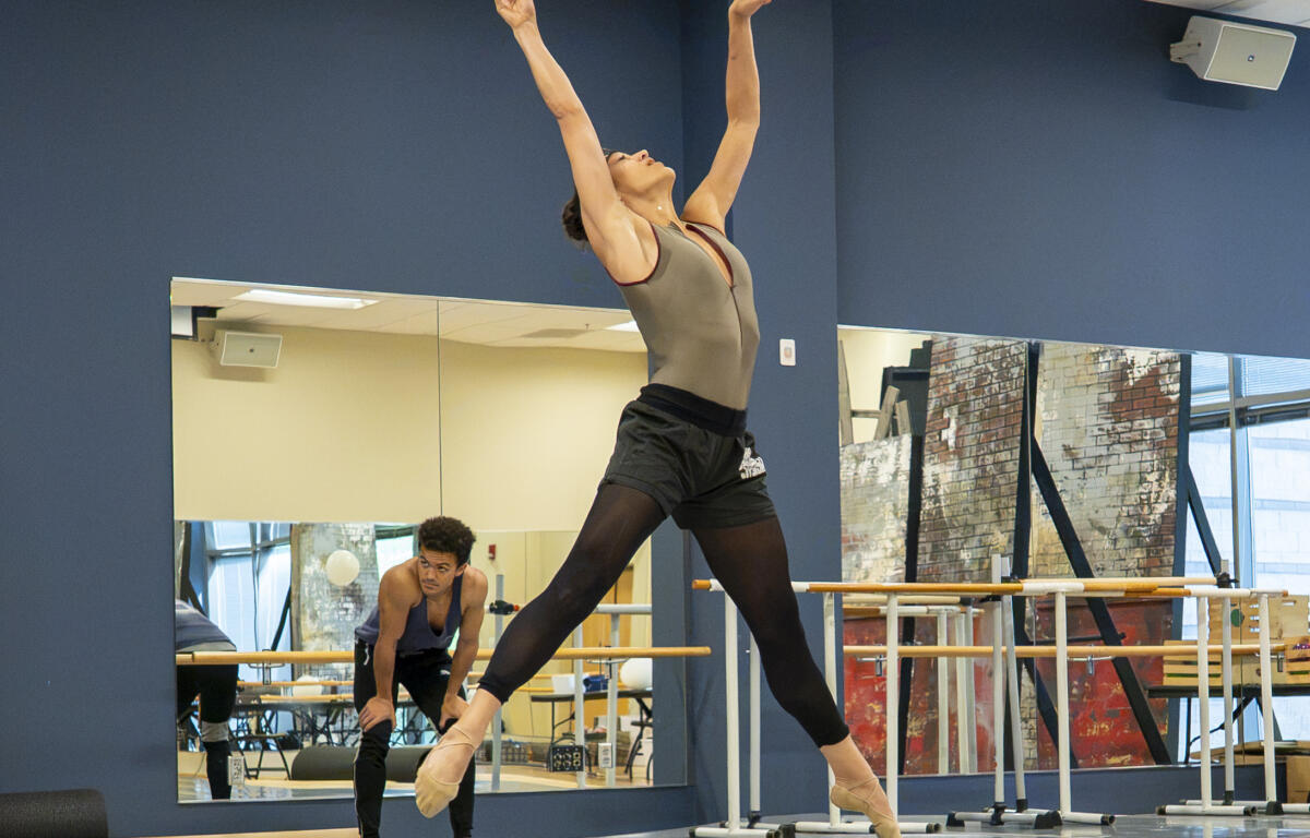Dancer leaps on pointe with arms raised in a mirrored dance studio; another dancer sits in the background by the barre.