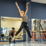 Dancer leaps on pointe with arms raised in a mirrored dance studio; another dancer sits in the background by the barre.