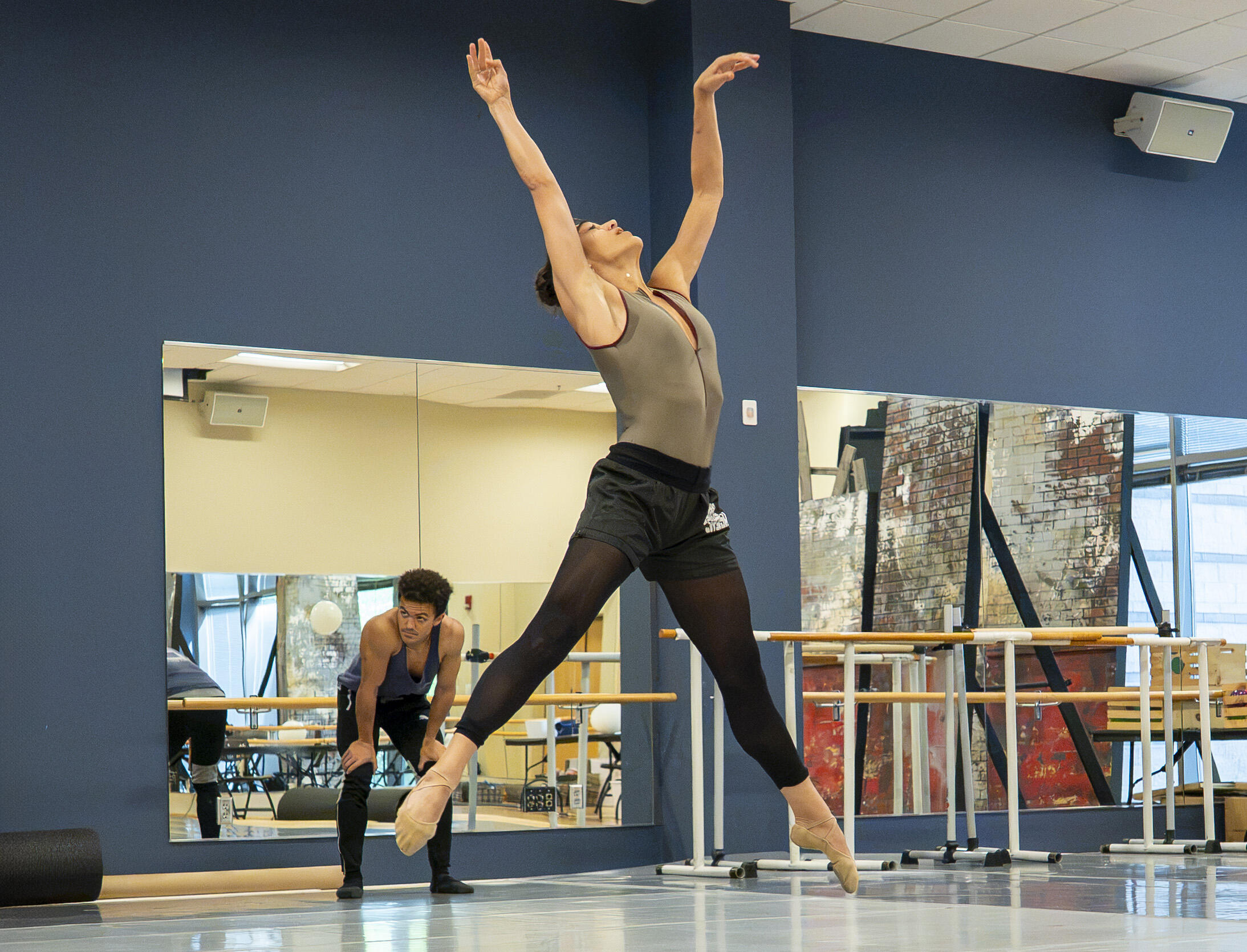 Dancer leaps on pointe with arms raised in a mirrored dance studio; another dancer sits in the background by the barre.