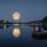 Fireworks over a calm lake at dusk, reflected in the water, while three people sit on a wooden dock watching.