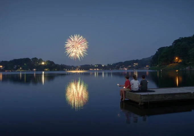 Fireworks over a calm lake at dusk, reflected in the water, while three people sit on a wooden dock watching.