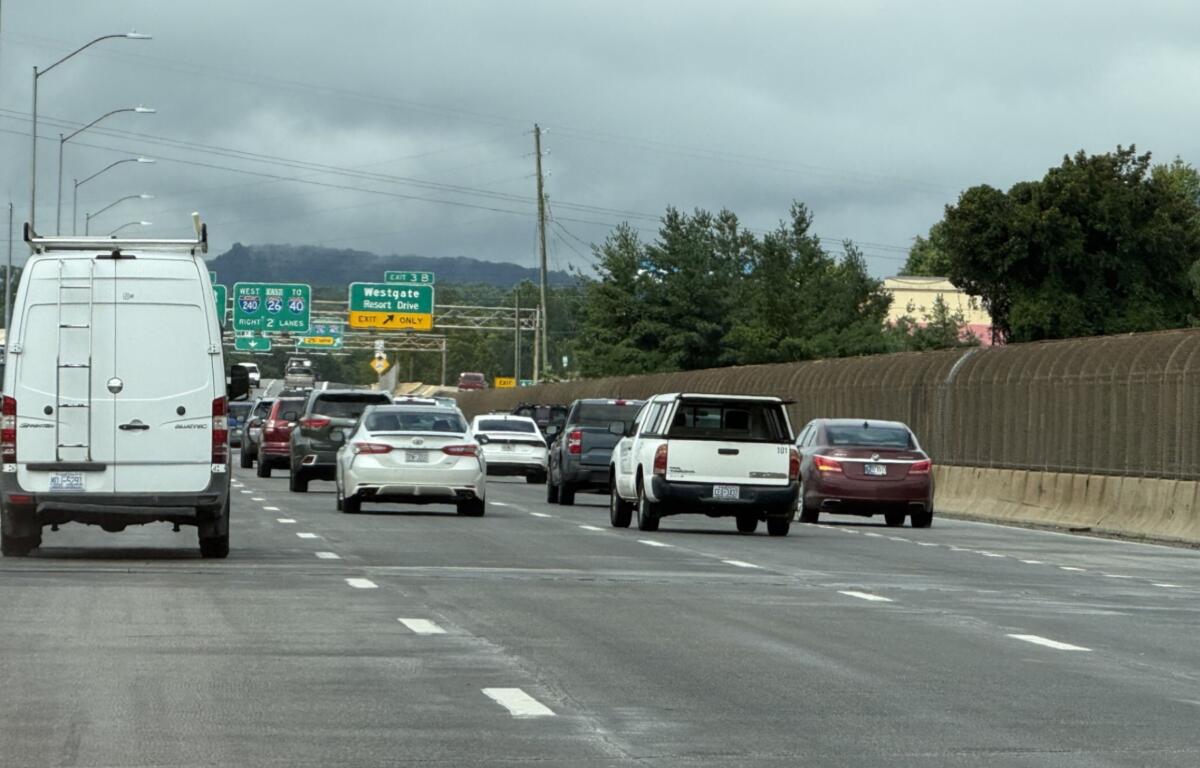 Busy multi-lane freeway scene with a white van in the foreground and numerous cars ahead under an overcast sky. Signage visible in the distance.