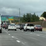 Busy multi-lane freeway scene with a white van in the foreground and numerous cars ahead under an overcast sky. Signage visible in the distance.