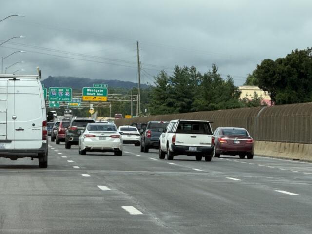 Busy multi-lane freeway scene with a white van in the foreground and numerous cars ahead under an overcast sky. Signage visible in the distance.