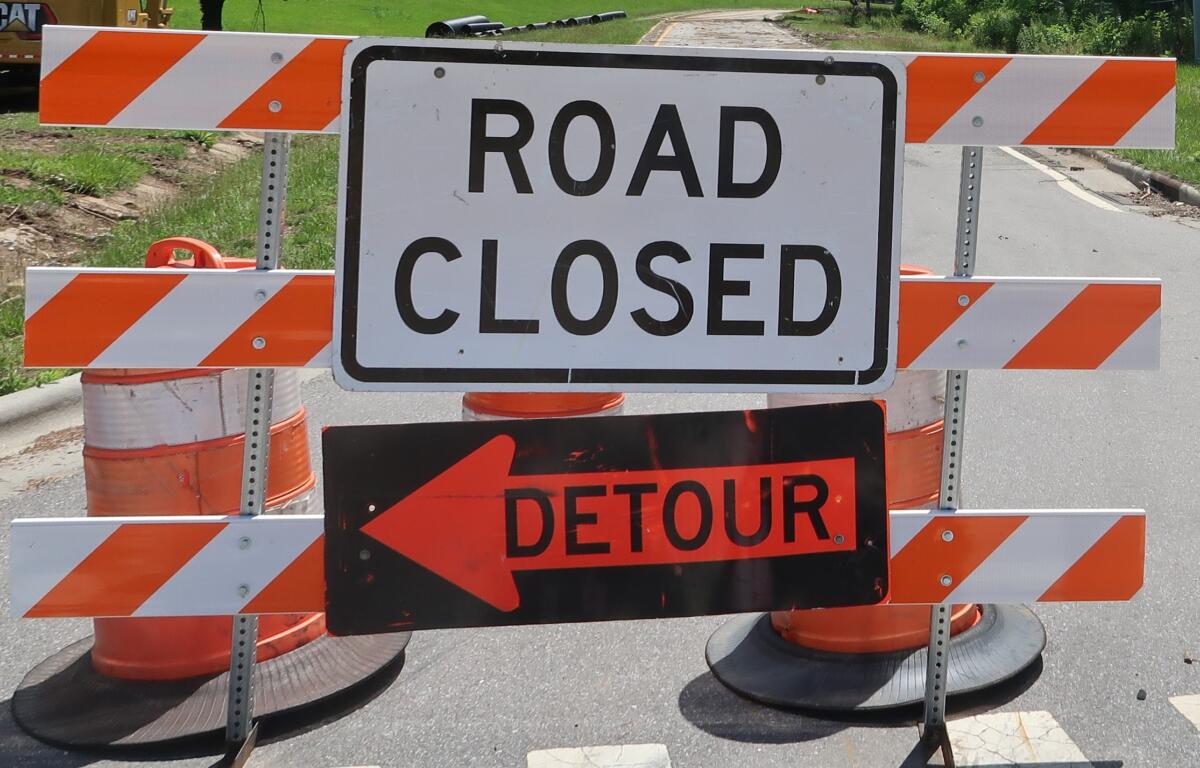 Road closed barrier with orange-and-white striped barricades and a detour sign blocking the road, directing traffic away from the closed area.
