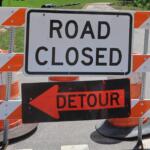 Road closed barrier with orange-and-white striped barricades and a detour sign blocking the road, directing traffic away from the closed area.