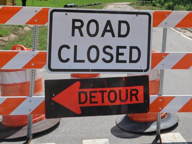 Road closed barrier with orange-and-white striped barricades and a detour sign blocking the road, directing traffic away from the closed area.