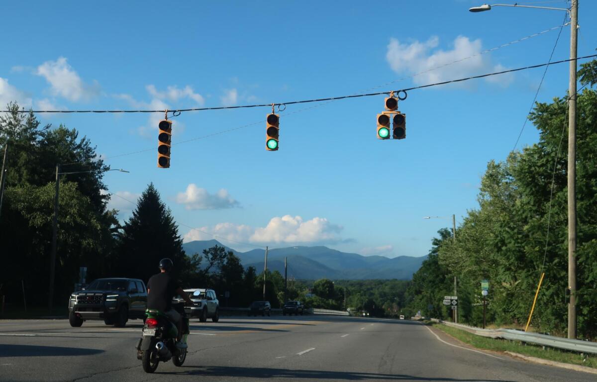 Green traffic lights above a multi-lane road with vehicles, including a foreground motorcycle, against a blue sky and distant mountains.