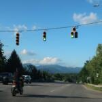 Green traffic lights above a multi-lane road with vehicles, including a foreground motorcycle, against a blue sky and distant mountains.