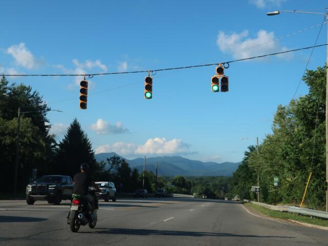 Green traffic lights above a multi-lane road with vehicles, including a foreground motorcycle, against a blue sky and distant mountains.