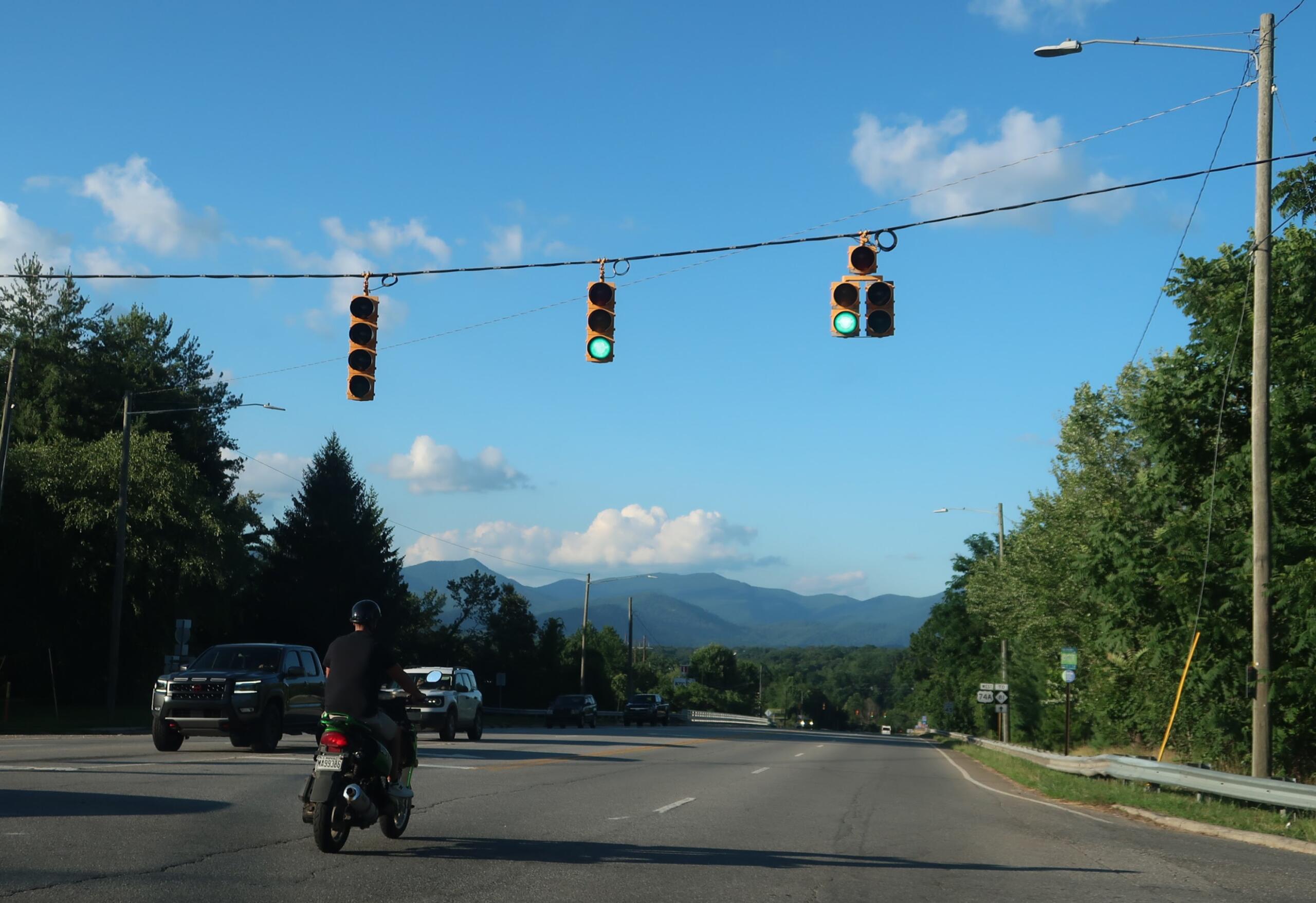 Green traffic lights above a multi-lane road with vehicles, including a foreground motorcycle, against a blue sky and distant mountains.