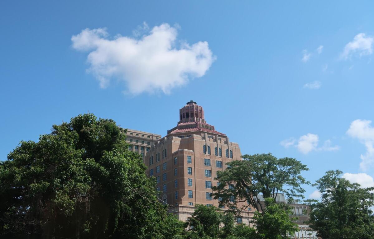 Tall beige brick building with a stepped red-tile crown rises behind green trees under a clear blue sky with a small white cloud nearby.