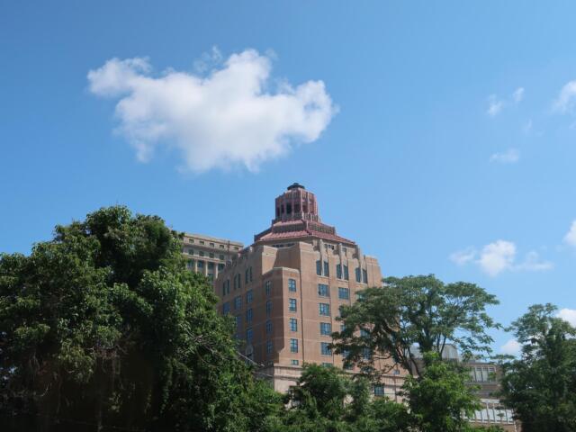 Tall beige brick building with a stepped red-tile crown rises behind green trees under a clear blue sky with a small white cloud nearby.