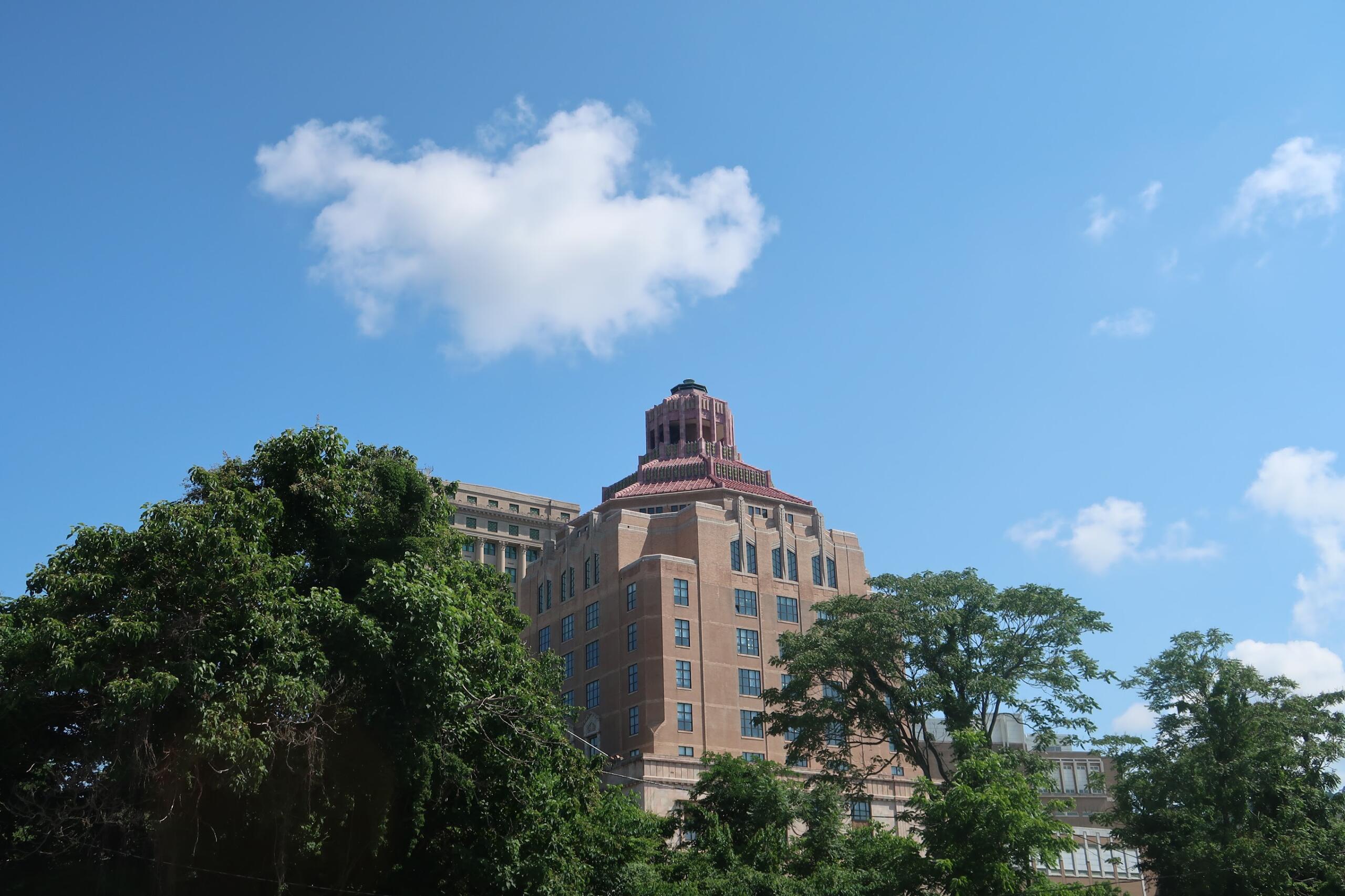 Tall beige brick building with a stepped red-tile crown rises behind green trees under a clear blue sky with a small white cloud nearby.