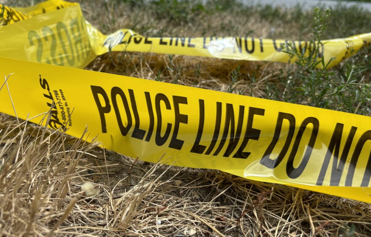 Yellow police tape reading 'POLICE LINE DO NOT CROSS' stretched across dry grass in an outdoor area, signaling a restricted scene.
