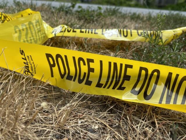 Yellow police tape reading 'POLICE LINE DO NOT CROSS' stretched across dry grass in an outdoor area, signaling a restricted scene.