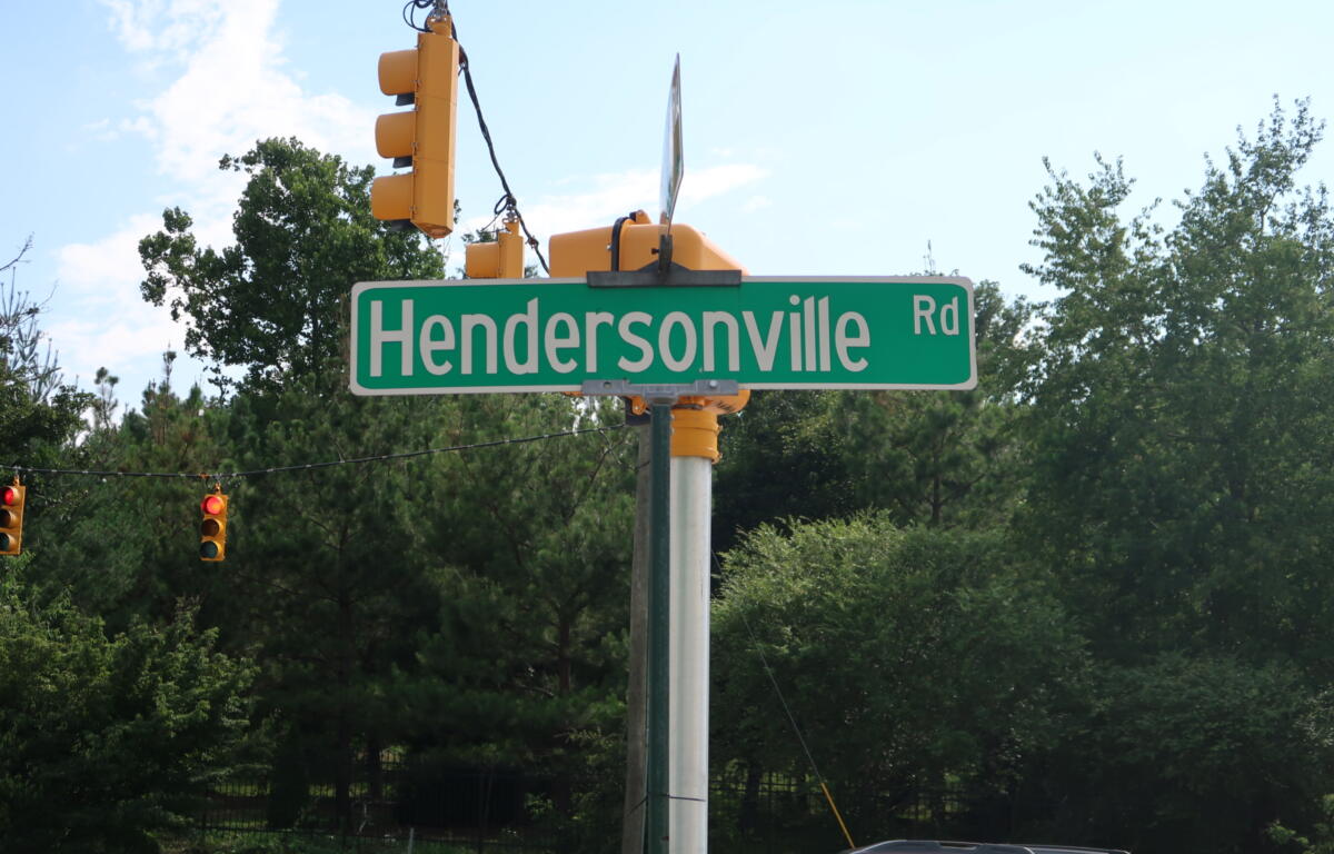Green Hendersonville Rd street sign on a pole with yellow traffic lights overhead and blue sky behind.