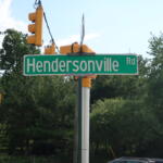 Green Hendersonville Rd street sign on a pole with yellow traffic lights overhead and blue sky behind.