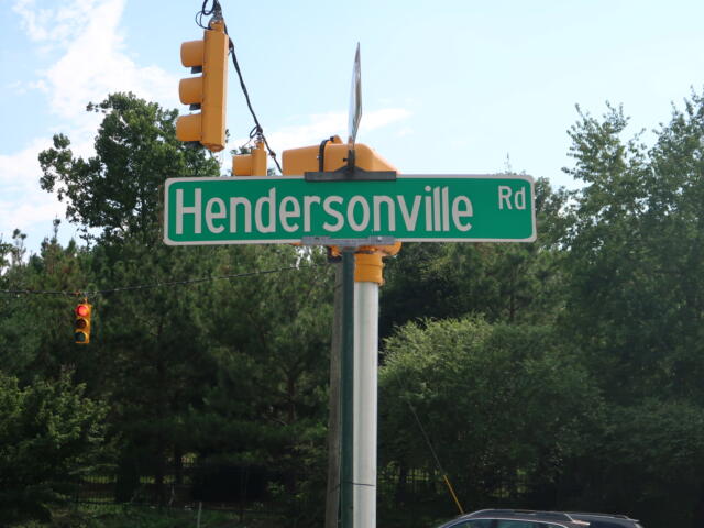 Green Hendersonville Rd street sign on a pole with yellow traffic lights overhead and blue sky behind.