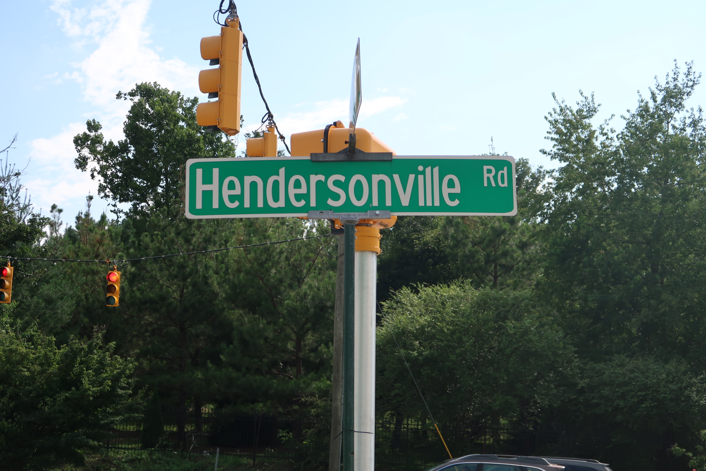 Green Hendersonville Rd street sign on a pole with yellow traffic lights overhead and blue sky behind.