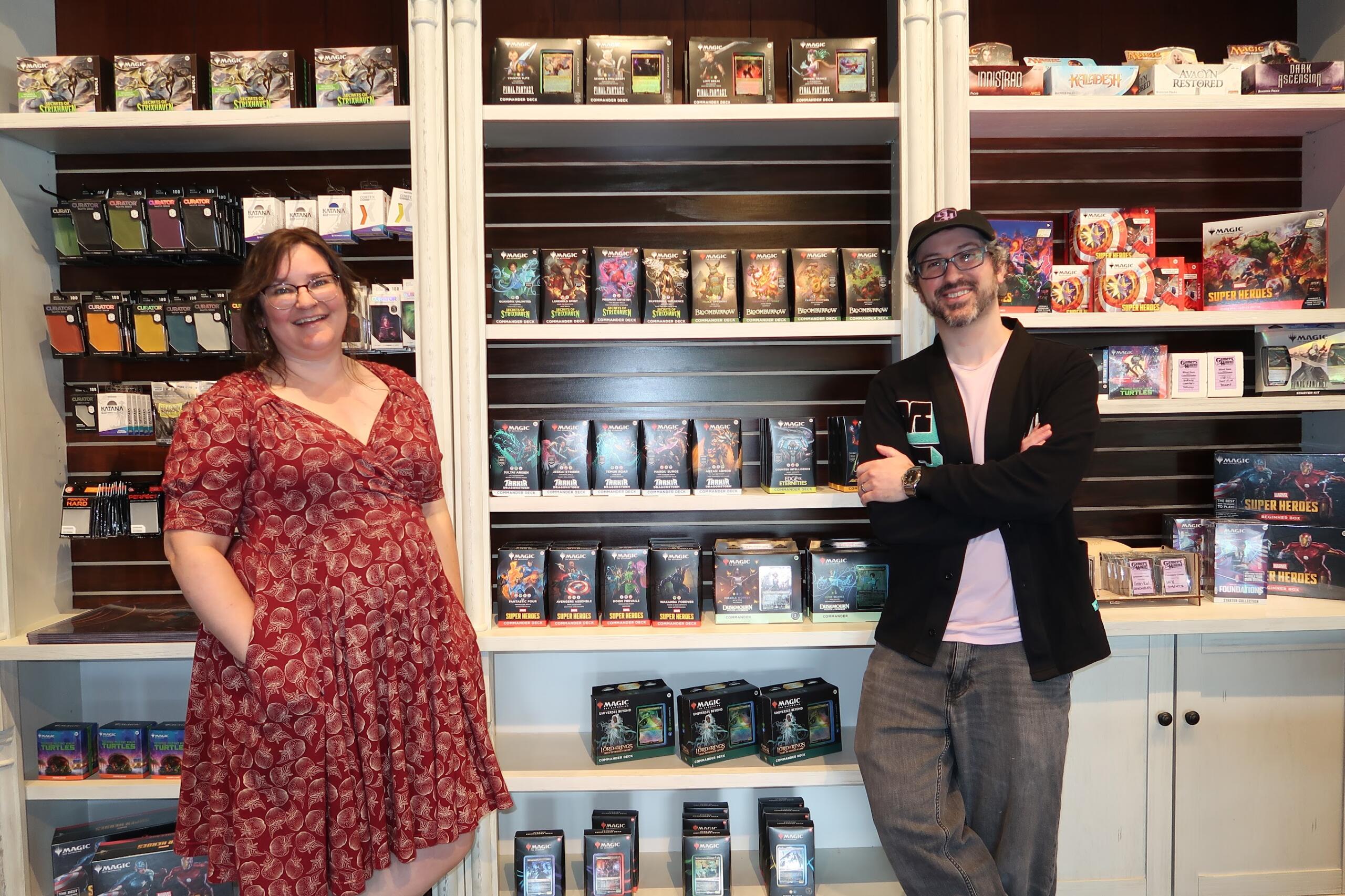 Two people stand in a game store in front of shelves filled with Magic: The Gathering card packs, smiling at the camera.