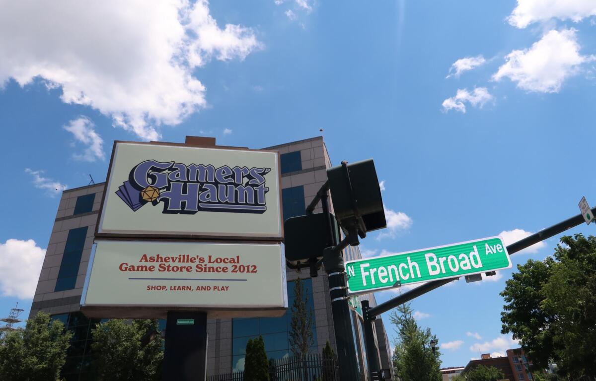 Sign for Gamers Haunt, Asheville's local game store since 2012, under a bright blue sky with clouds; street signs for French Broad Ave are visible in the foreground.