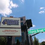 Sign for Gamers Haunt, Asheville's local game store since 2012, under a bright blue sky with clouds; street signs for French Broad Ave are visible in the foreground.