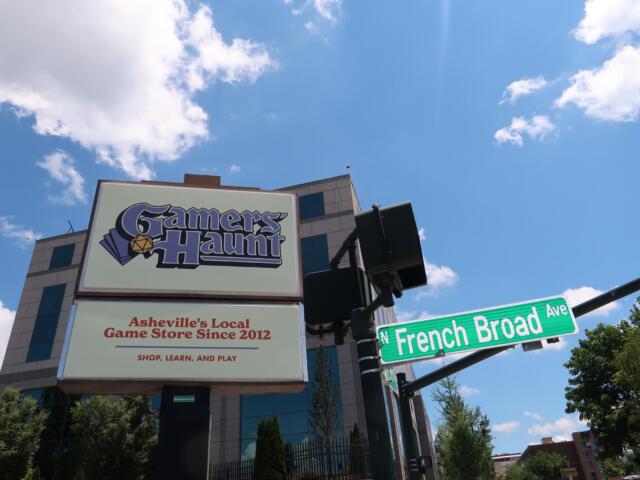 Sign for Gamers Haunt, Asheville's local game store since 2012, under a bright blue sky with clouds; street signs for French Broad Ave are visible in the foreground.
