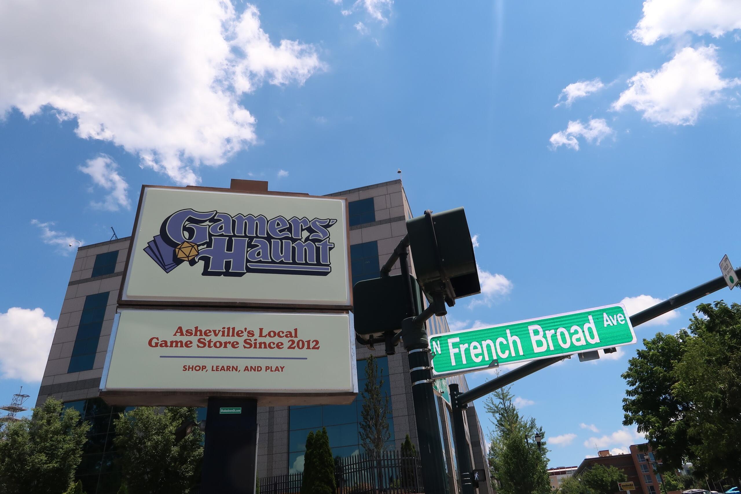 Sign for Gamers Haunt, Asheville's local game store since 2012, under a bright blue sky with clouds; street signs for French Broad Ave are visible in the foreground.