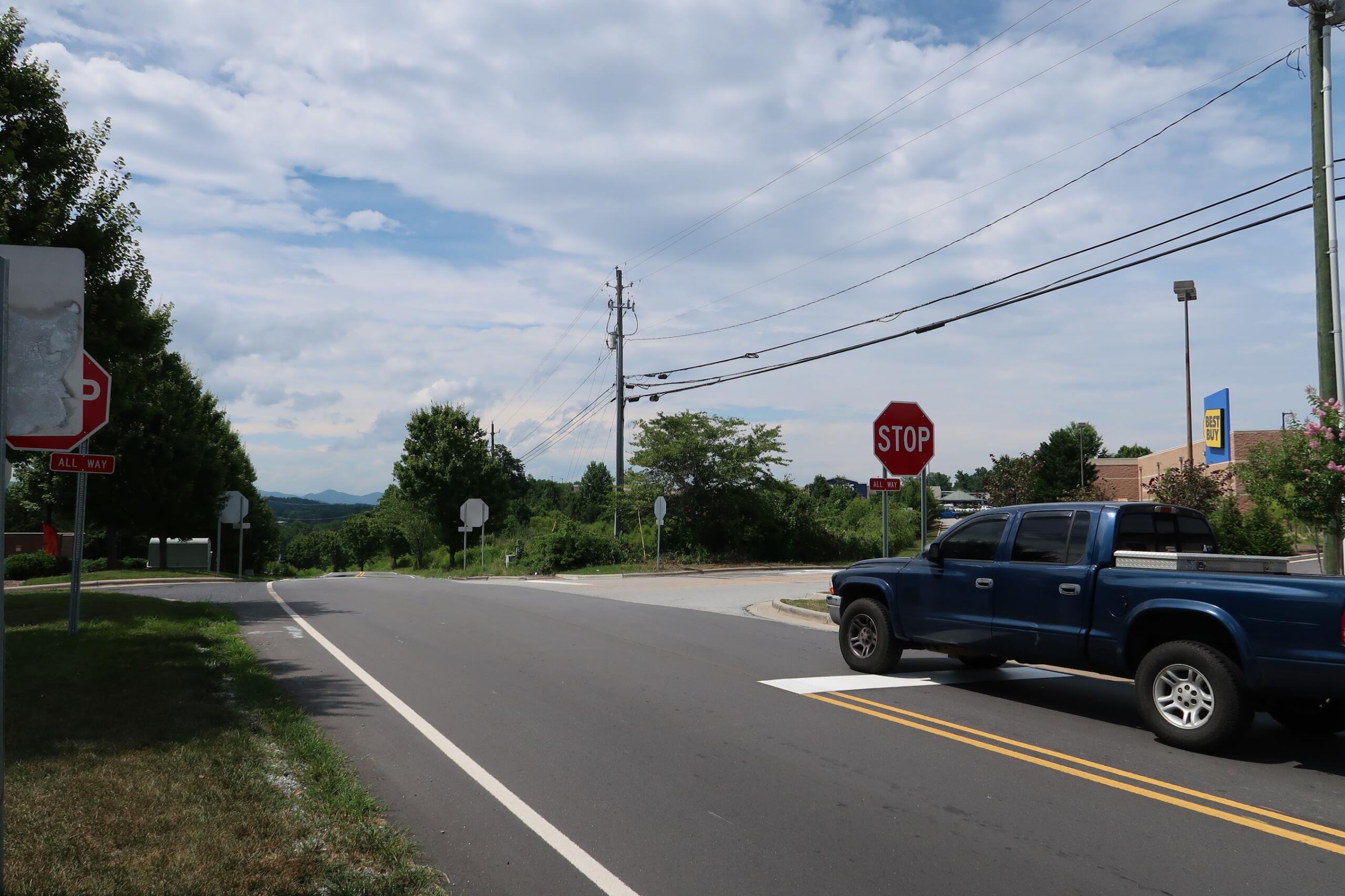 Suburban street intersection with stop signs, a blue pickup truck in the foreground, and power lines overhead against a cloudy sky